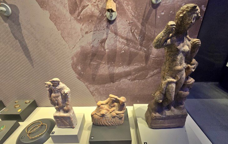 İznik Müzesi’nde sergilenen Roma dönemine ait kadın figürü, mitolojik sahneli küçük heykelcikler ve mermer betimlemelerin birlikte görüldüğü vitrin düzenlemesi.